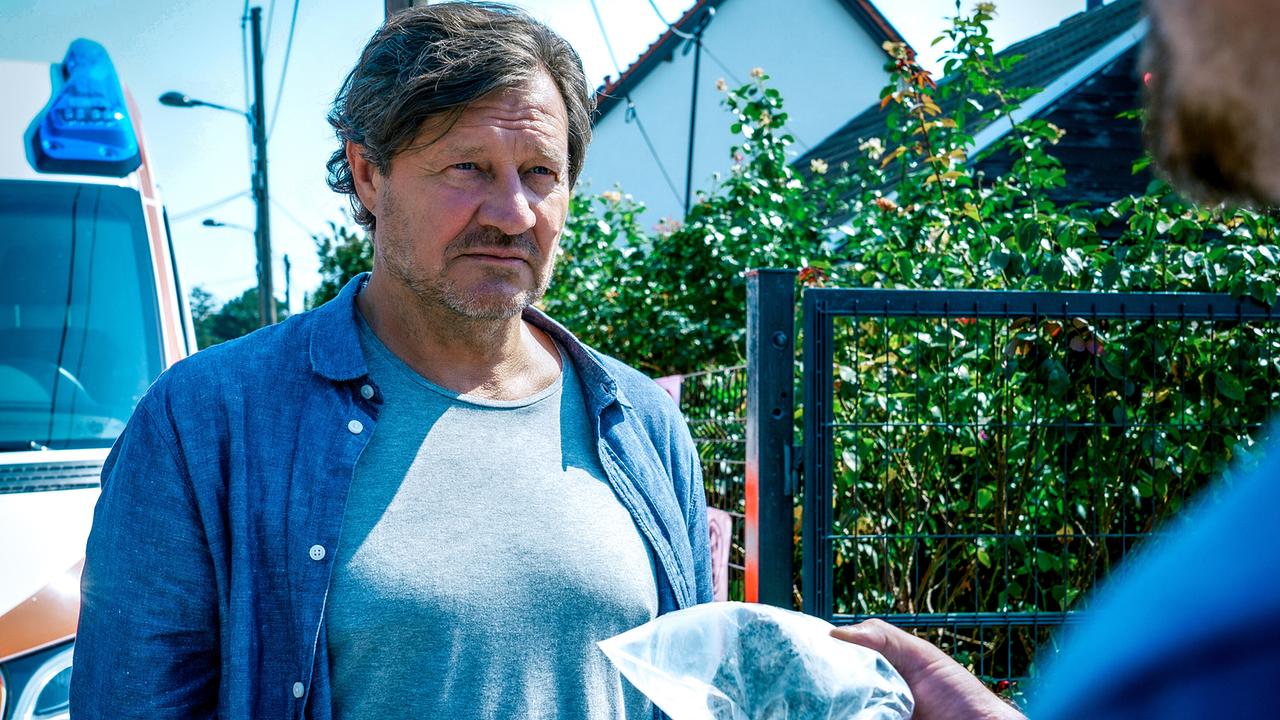 © ORF/Satel Film/Petro Domenigg Vor einem Gartentor und einem Rettungswagen steht Gernot Polmer, im Vordergrund ist unscharf Max Herzog und jemand, der einen durchsichtigen Beutel hält, zu erkennen.