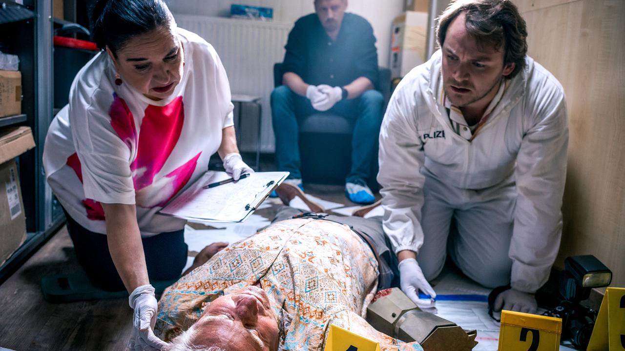 © ORF/Satel Film/Luca Breuer Franziska Beck und Julius Rubatsch knien am Boden und untersuchen den am Boden liegende Toten Helmut Brandl in einem Innenraum mit markierten Beweismitteln. Im Hintergrund sitzt Max Herzog.