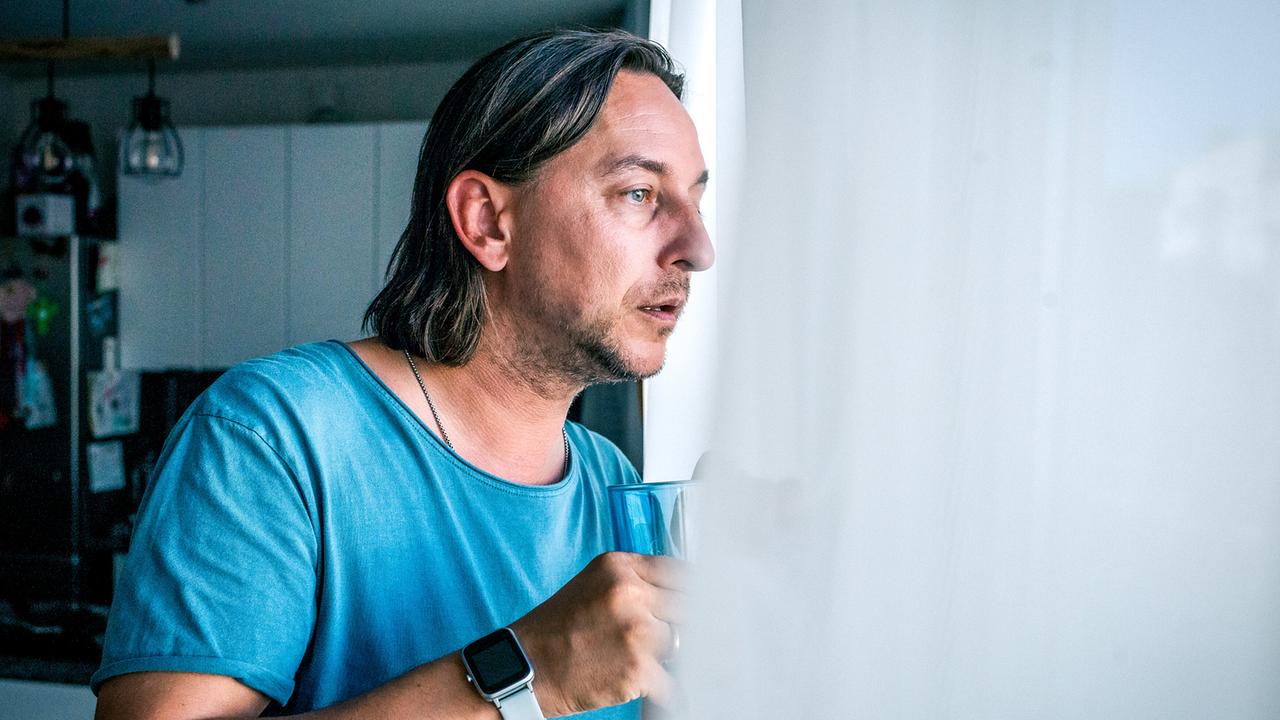 © ORF/Satel Film/Luca Breuer Christian Denk steht in einem hellen Wohnraum am Fenster, hält eine Tasse in der Hand und schaut hinaus.