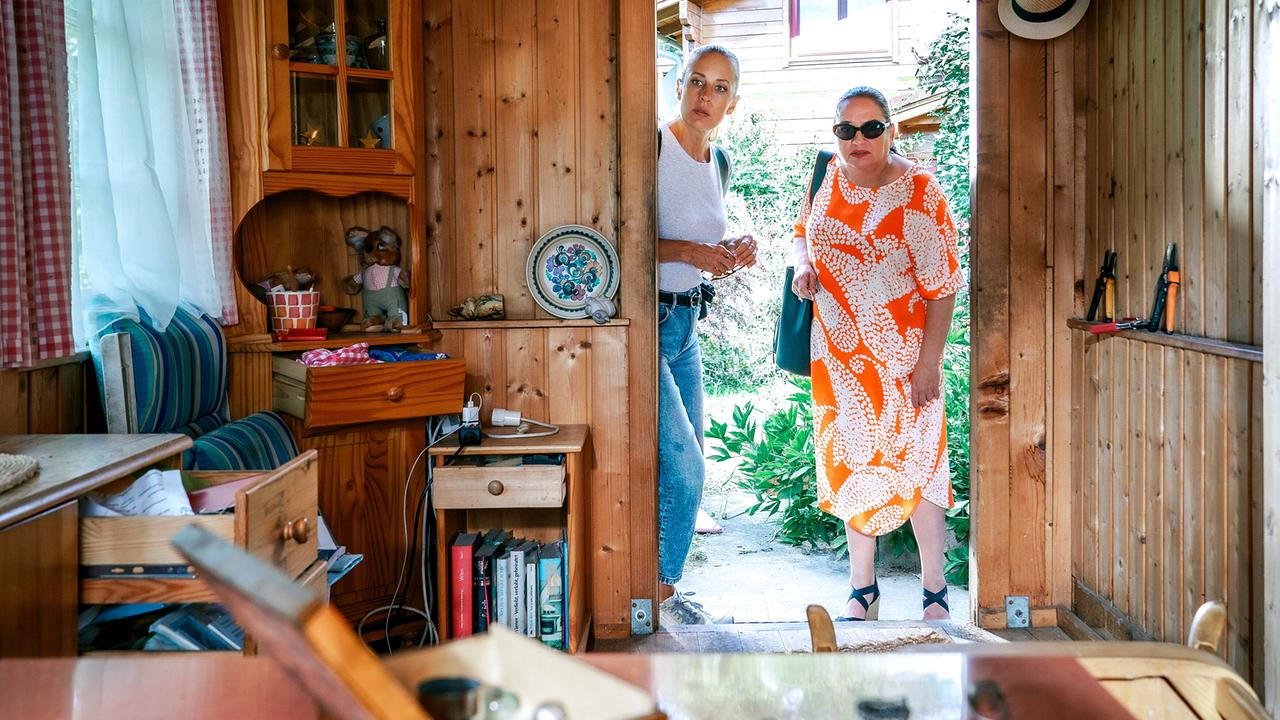 © ORF/Satel Film/Petro Domenigg Penny Lanz undFranziska Beck stehen in der Tür einer rustikalen Holzhütte und blicken in den Innenraum mit Möbeln, offenen Schubladen und Alltagsgegenständen.