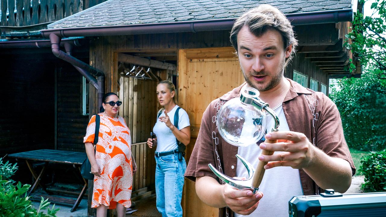 © ORF/Satel Film/Petro Domenigg Julius Rubatsch begutachtet im Vordergrund ein Glasobjekt in einem Schrebergarten, während Franziska Beck und Penny Lanz vor einer Holzhütte im Hintergrund stehen.