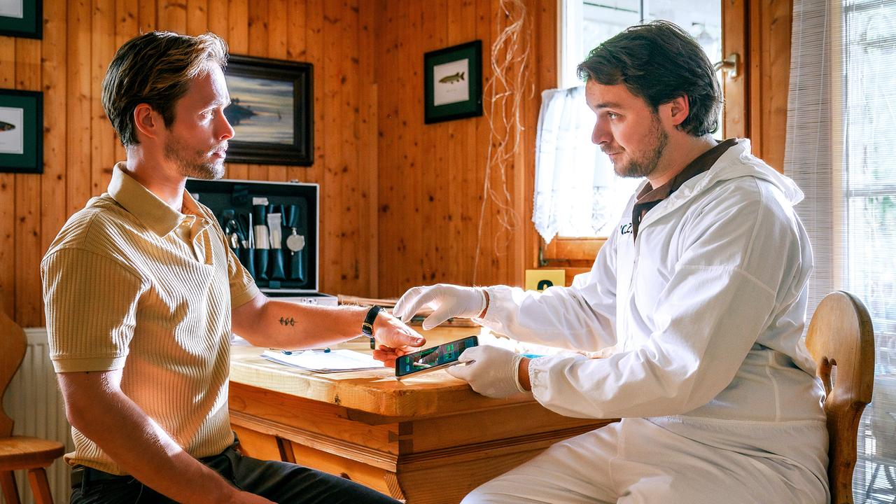 © ORF/Satel Film/Petro Domenigg In einem Holzraum sitzen Simon Rossi und Julius Rubatsch mit Schutzkleidung an einem Tisch. Julius hält Simon ein Smartphone hin, dessen Daumen knapp über dem Bildschirm liegt.