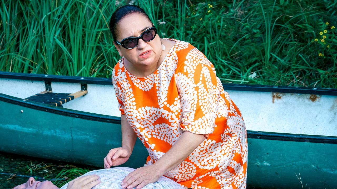 © ORF/Satel Film/Petro Domenigg Franziska Beck in einem orange-weißen Kleid und schwarzer Sonnenbrille kniet neben einem am Boden liegenden Mann in der Nähe eines Kanus.