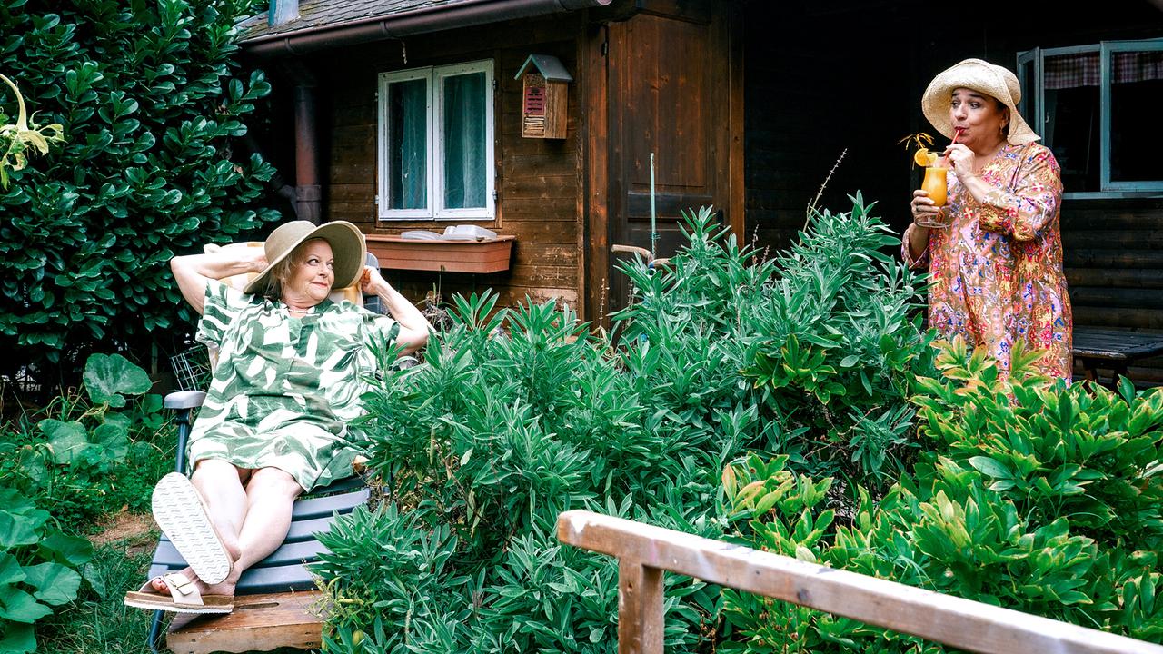 © ORF/Satel Film/Petro Domenigg Henriette Wolf liegt in einem Schrebergarten vor einer Holzhütte auf einer Gartenliege. Ein paar Meter neben ihr steht Franziska Beck und trinkt einen orangefarbenen Cocktail mit einem Strohhalm.