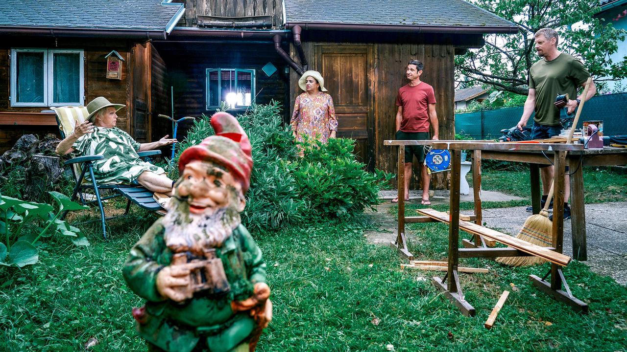© ORF/Satel Film/Petro Domenigg Vor einem Holzhaus liegt Henriette Wolf in einer Gartenliege, daneben stehen Franziska Beck sowie Klaus Lechner mit einer Kabeltrommel und Max Herzog vor einem Arbeitstisch. Im Vordergrund steht ein Gartenzwerg.