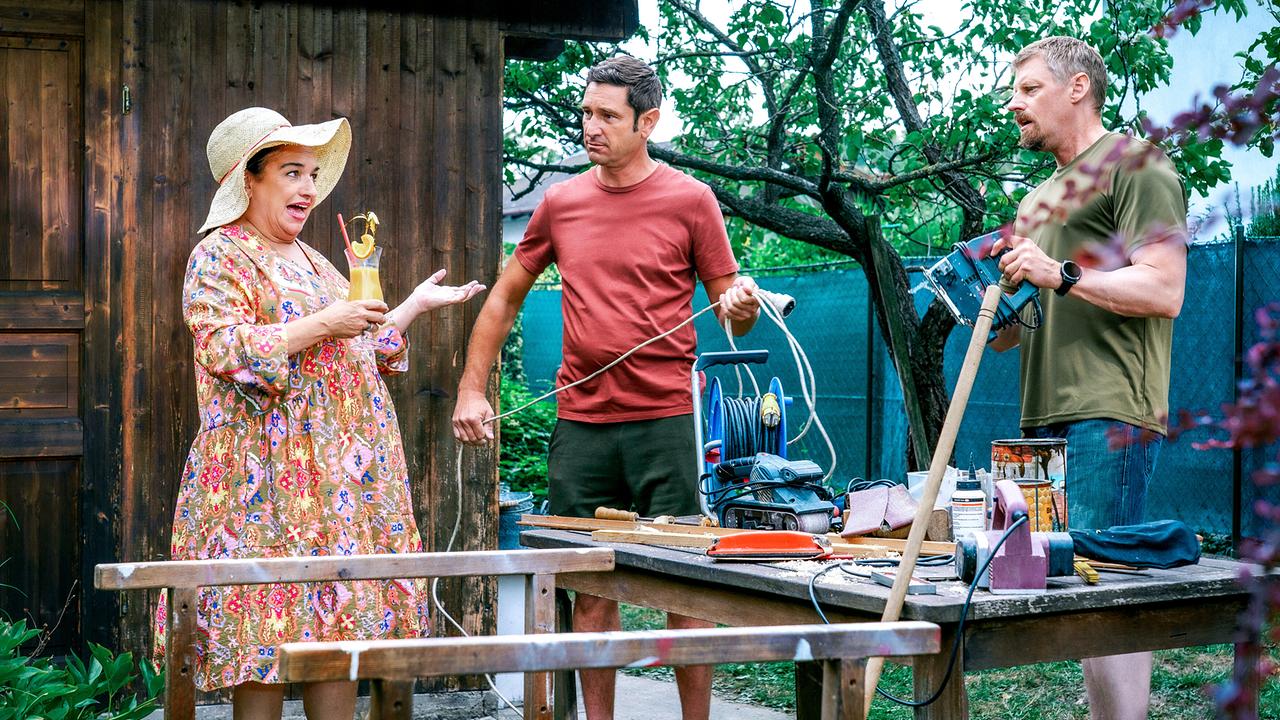 © ORF/Satel Film/Petro Domenigg Franziska Beck mit einem Cocktail in der Hand steht Klaus Lechner und Max Herzog, die an einem Arbeitstisch im Garten mit Werkzeugen und Kabeln hantieren.