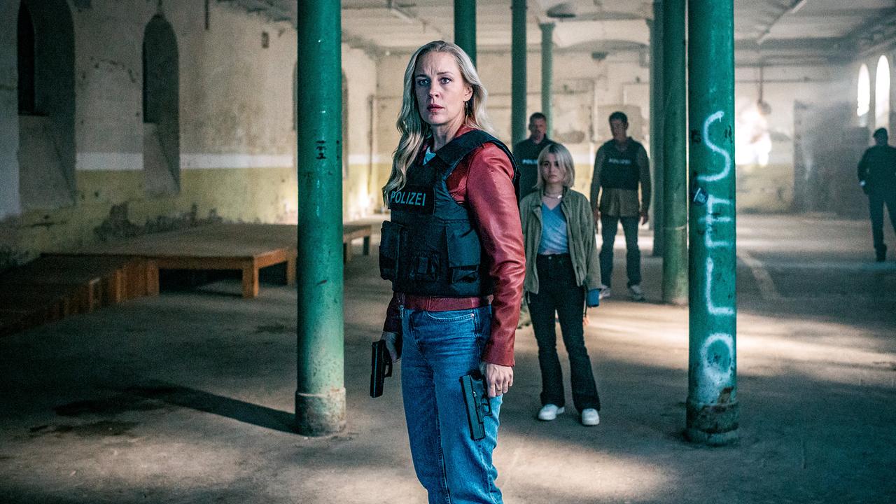 Penny Lanz mit schusssicherer Weste steht mit gezogener Waffe in einer verlassenen Industriehalle, begleitet von Max Herzog, Lea Schlatterer und Klaus Lechner im Hintergrund.