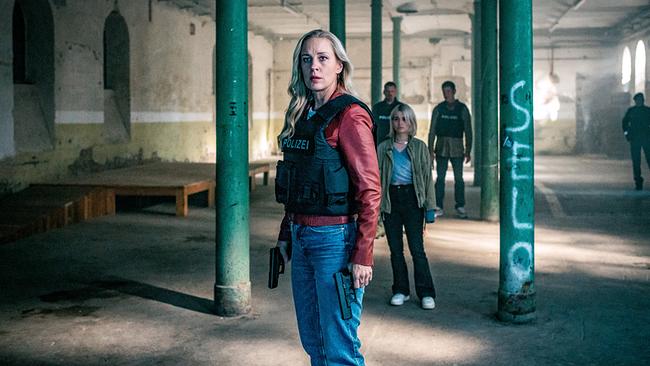 Penny Lanz mit schusssicherer Weste steht mit gezogener Waffe in einer verlassenen Industriehalle, begleitet von Max Herzog, Lea Schlatterer und Klaus Lechner im Hintergrund.