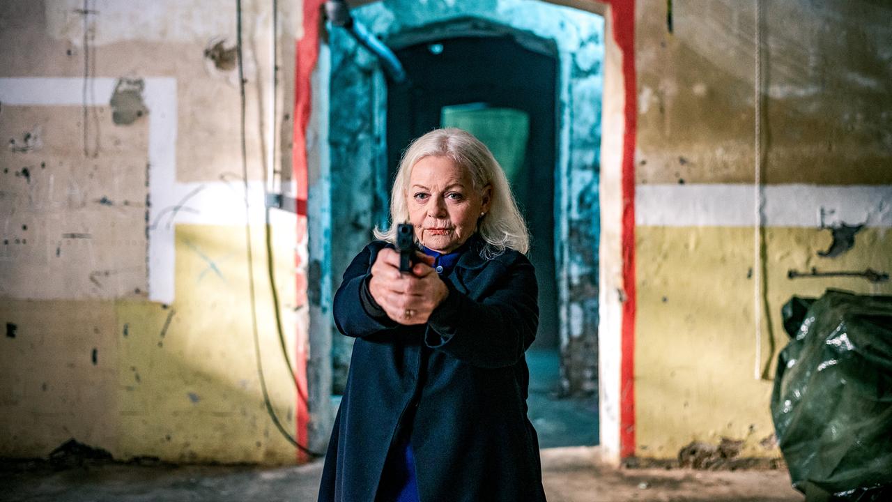 Henriette Wolf steht in einem verfallenem Raum und richtet eine Pistole direkt nach vorn.