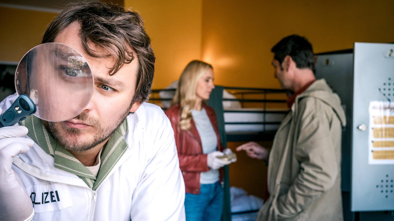Forensiker Julius Rubatsch mit Lupe steht in einem Zimmer mit Stockbetten im Vordergrund, während Penny Lanz und Klaus Lechner im Hintergrund etwas begutachten.
