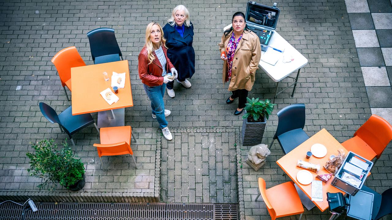 Eine Draufsicht zeigt Penny Lanz, Henriette Wolf und Franziska Beck in einem Innenhof zwischen Tischen und Sesseln, während am unteren Bildrand ein markierter Tatortbereich zu sehen ist.