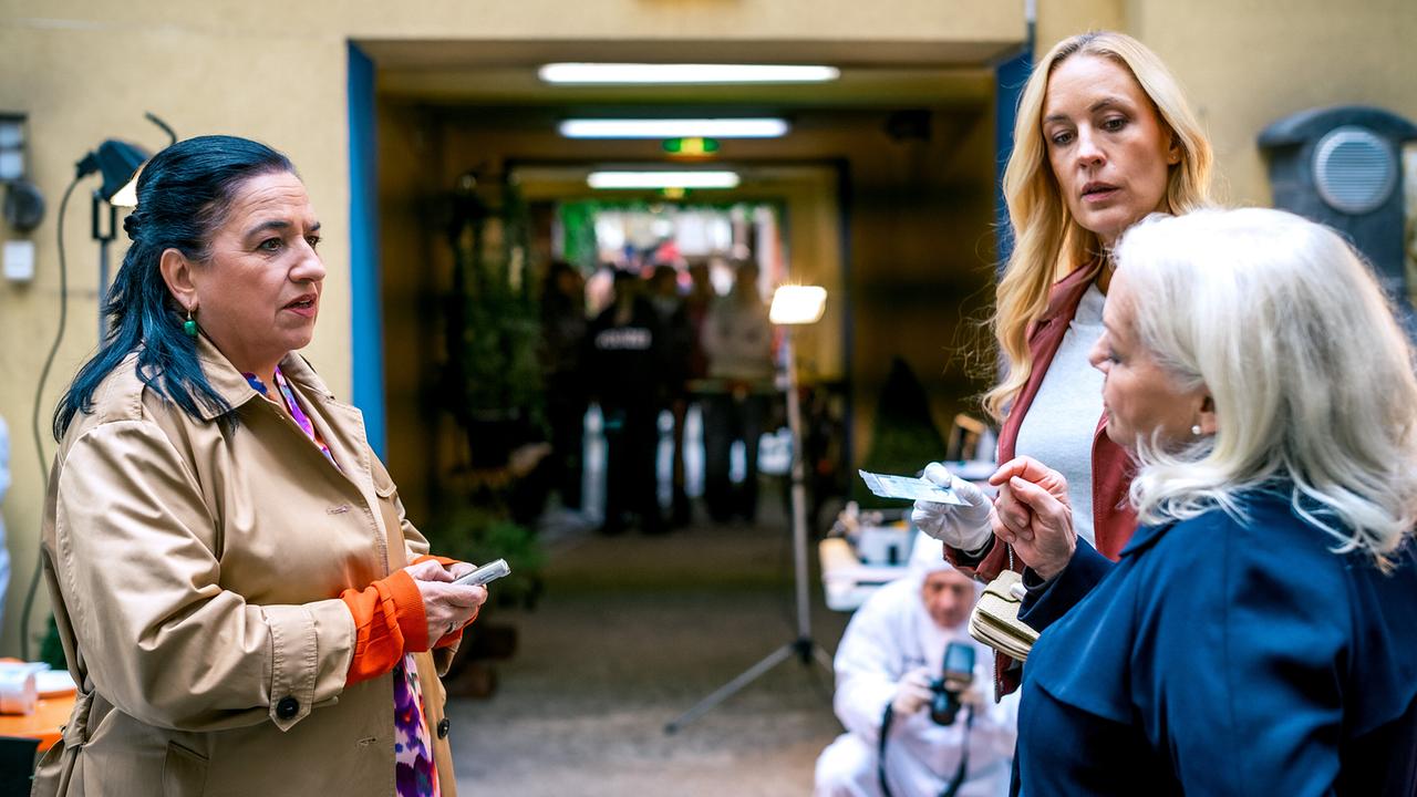 Franziska Beck, Penny Lanz und Henriette Wolf sprechen am Tatort vor einem Durchgang eines Innenhofs, während im Hintergrund weitere Ermittler zu sehen sind.