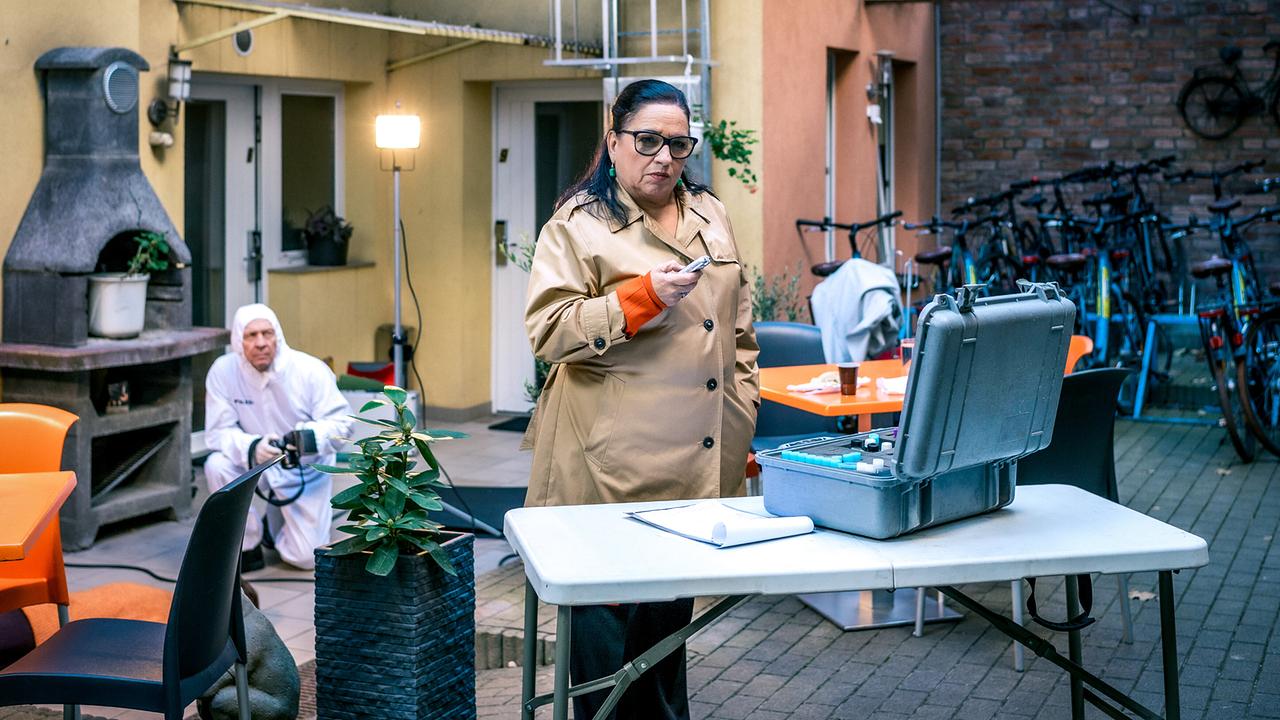 Franziska Beck im beigefarbenen Mantel steht in einem Hof bei einer kriminaltechnischen Untersuchung, während ein Forensiker im Hintergrund arbeitet.