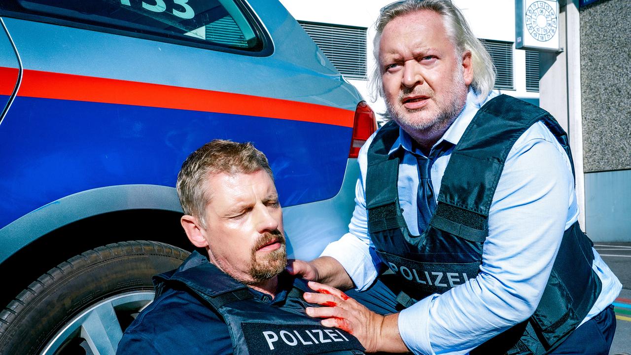 © ORF/Satel Film Max Herzog sitzt verletzt am Boden neben einem Einsatzfahrzeug, während Helmuth Nowak ihm Hilfe leistet.
