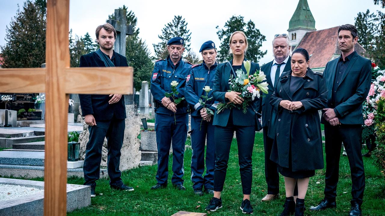 © ORF/Satel Film/Petro Domenigg Julius Rubatsch, Penny Lanz, Helmuth Nowak, Franziska Beck, Klaus Lechner und zwei uniformierte Polizisten stehen bei einer Beerdigung auf einem Friedhof vor einem offenen Grab mit einem Holzkreuz.