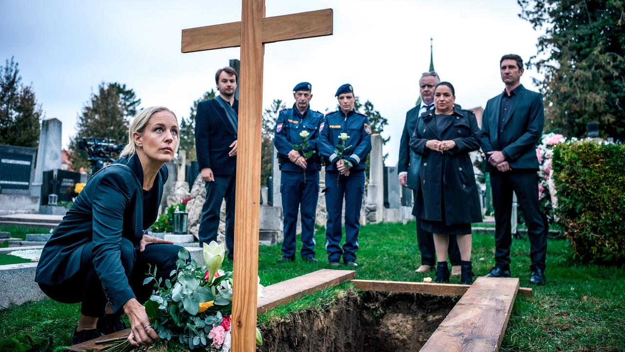 © ORF/Satel Film/Petro Domenigg Penny Lanz kniet an einem offenen Grab auf einem Friedhof und legt Blumen nieder, während mehrere Trauergäste im Hintergrund stehen.