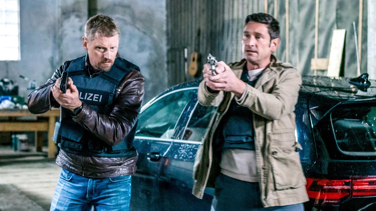 Max Herzog und Klaus Lechner, beide in Schutzwesten,  zielen in einer Industriehalle mit gezogenen Pistolen neben einem geparkten Auto nach vorne.