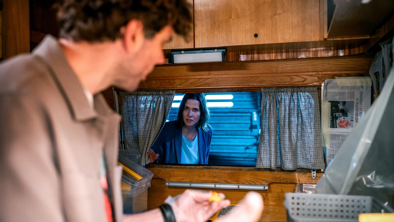 Vom Inneren des Wohnwagens, in dem Ron Wagner steht, aus ist durch ein Fenster Johanna Binder zu sehen, die von draußen hereinschaut.