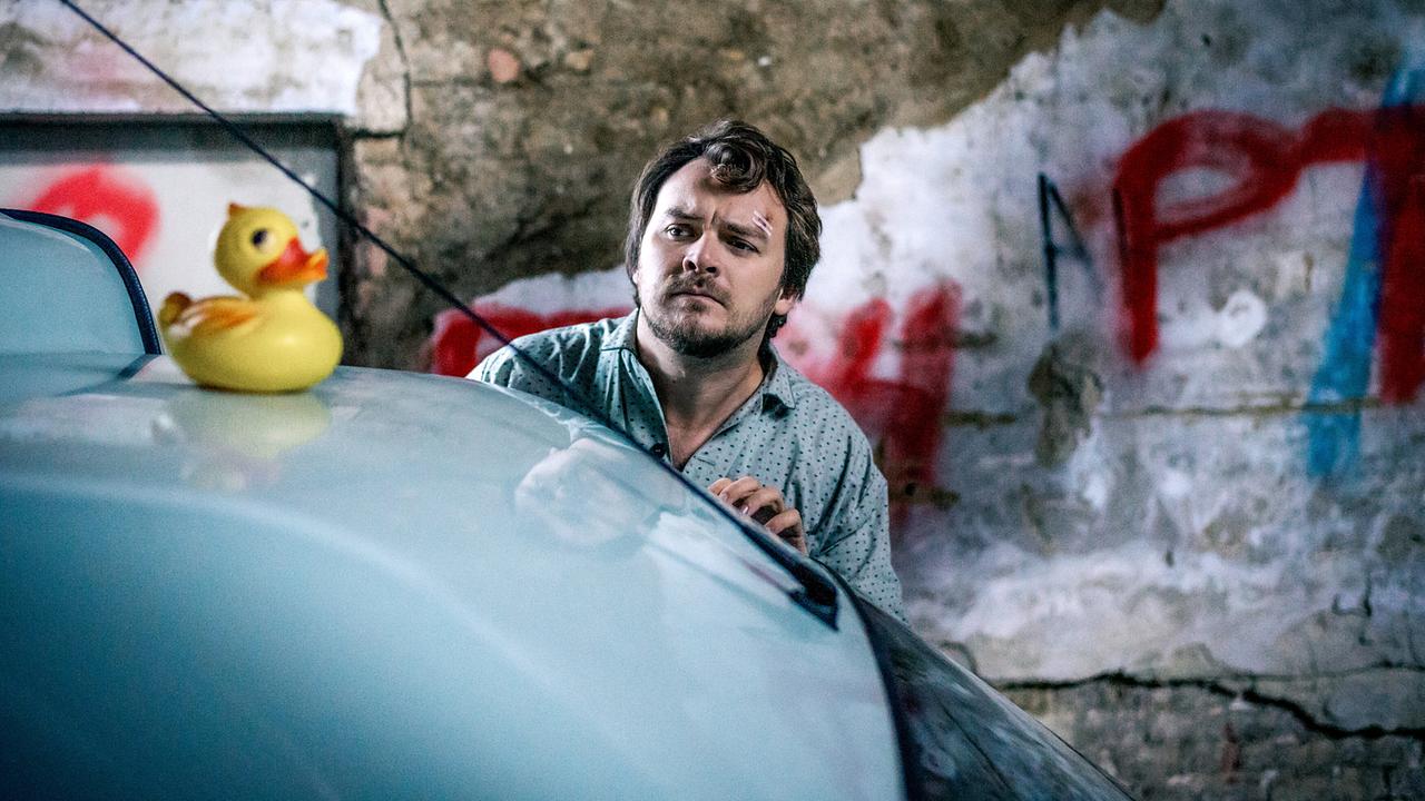 Julius Rubatsch lehnt sich über die Motorhaube eines Autos, auf der eine gelbe Gummiente steht, vor einer verwitterten, mit Graffiti besprühten Wand.