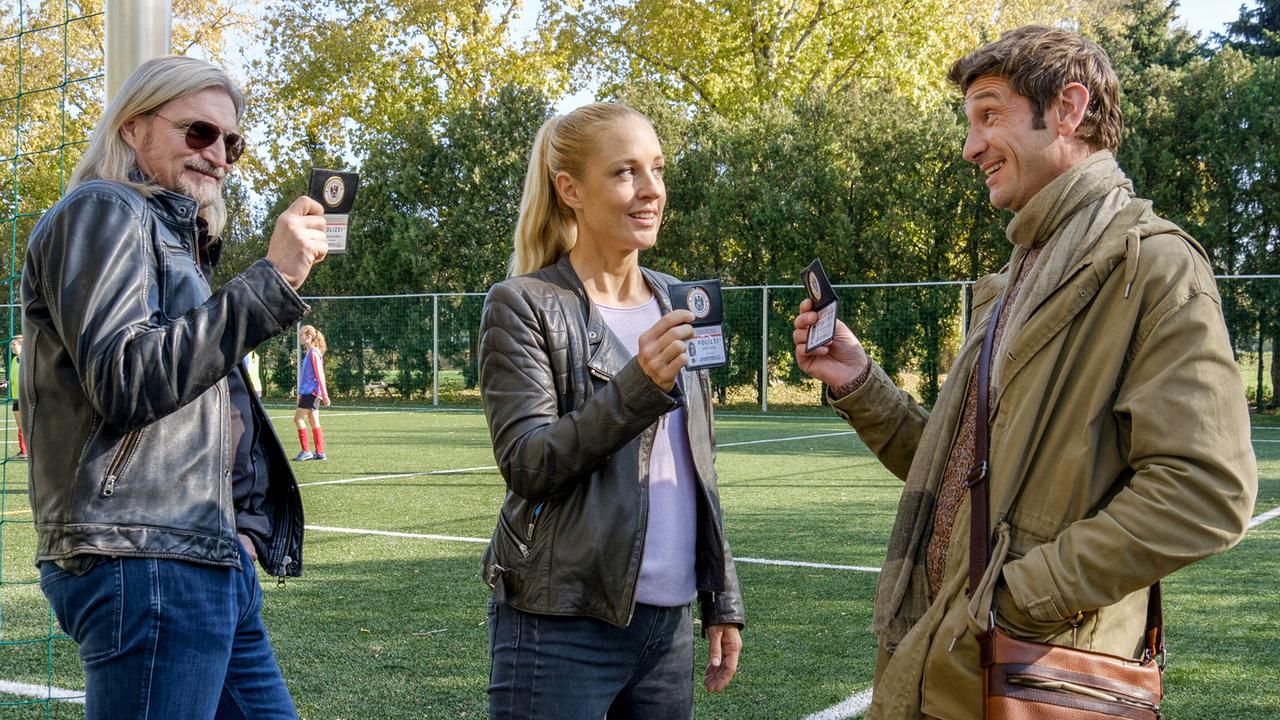 Carl, Penny und Klaus zeigen sich lächelnd untereinander ihre Ausweise am Rand eines Fußballfeldes.