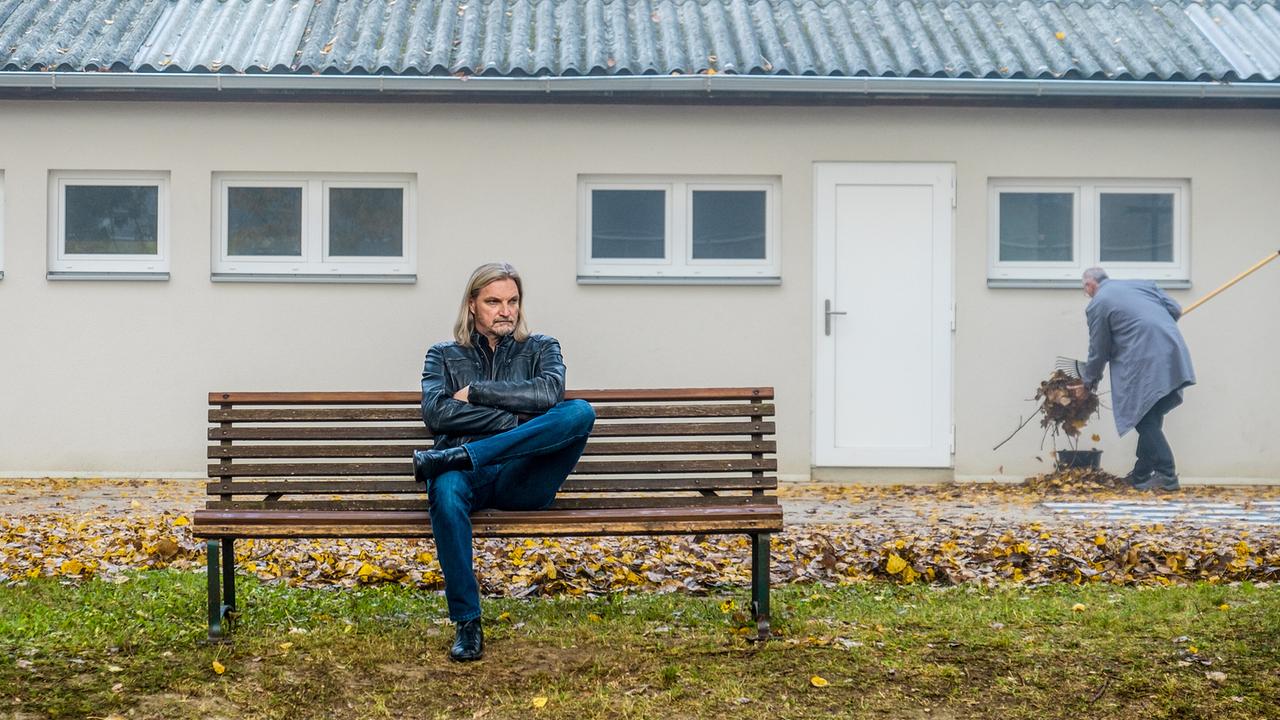 Carl Ribarski sitzt mit verschränkten Armen auf einer Parkbank vor einem Gebäude, im Hintergrund kehrt ein Mann Laub.