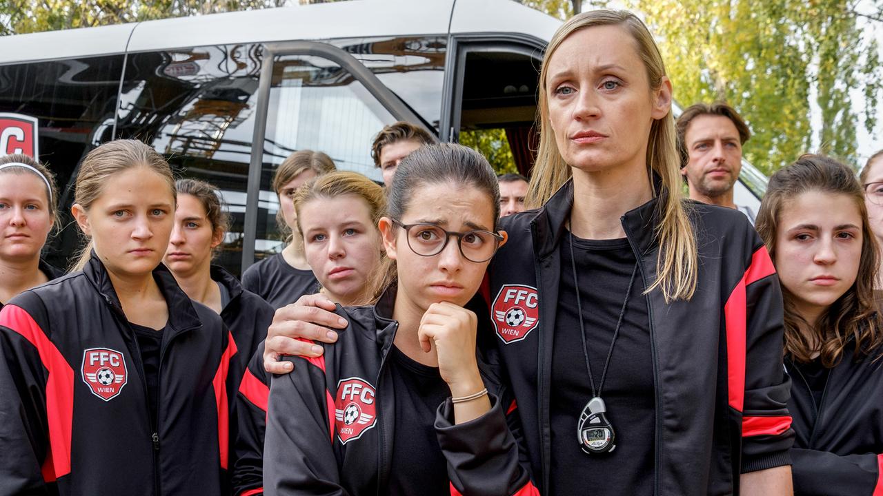 Eine Gruppe junger Fußballspielerinnen in Trainingsanzügen steht mit Trainerin Nina Hartl dicht beieinander vor einem Bus.
