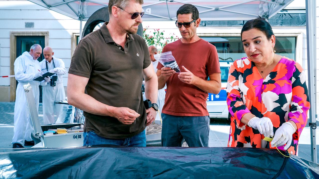 © ORF/Satel Film/Petro Domenigg Max Herzog, Klaus Lechner und Franziska Beck stehen am Tatort an einem Tisch unter einem Zelt und begutachten Beweismittel.