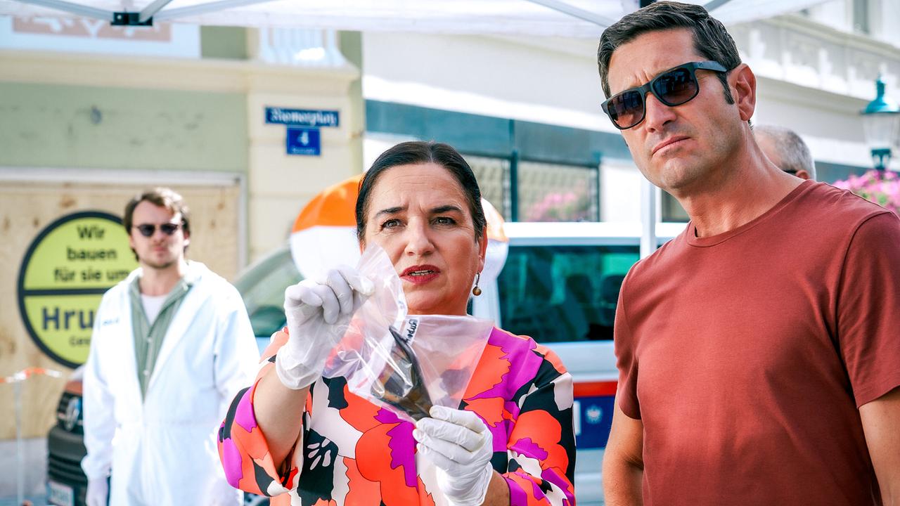 © ORF/Satel Film/Petro Domenigg Franziska Beck mit Handschuhen hält am Tatort im Freien einen Teil der Bierflasche in einem Plastikbeutel hoch, während Klaus Lechner danebensteht und Julius Rubatsch im Schutzanzug im Hintergrund zu sehen ist. .