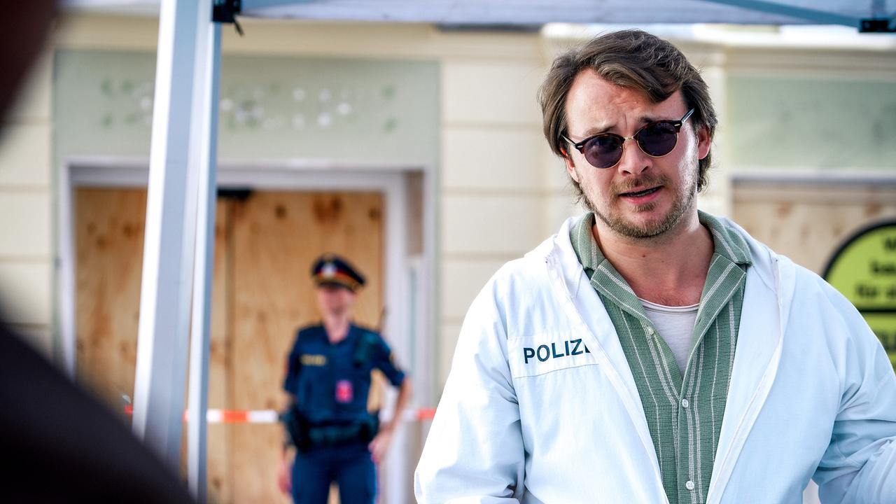 © ORF/Satel Film/Petro Domenigg Julius Rubatsch in einem weißen Schutzanzug mit der Aufschrift „Polizei“ steht vor einem abgesperrten Bereich im Freien, im Hintergrund eine Polizistin.