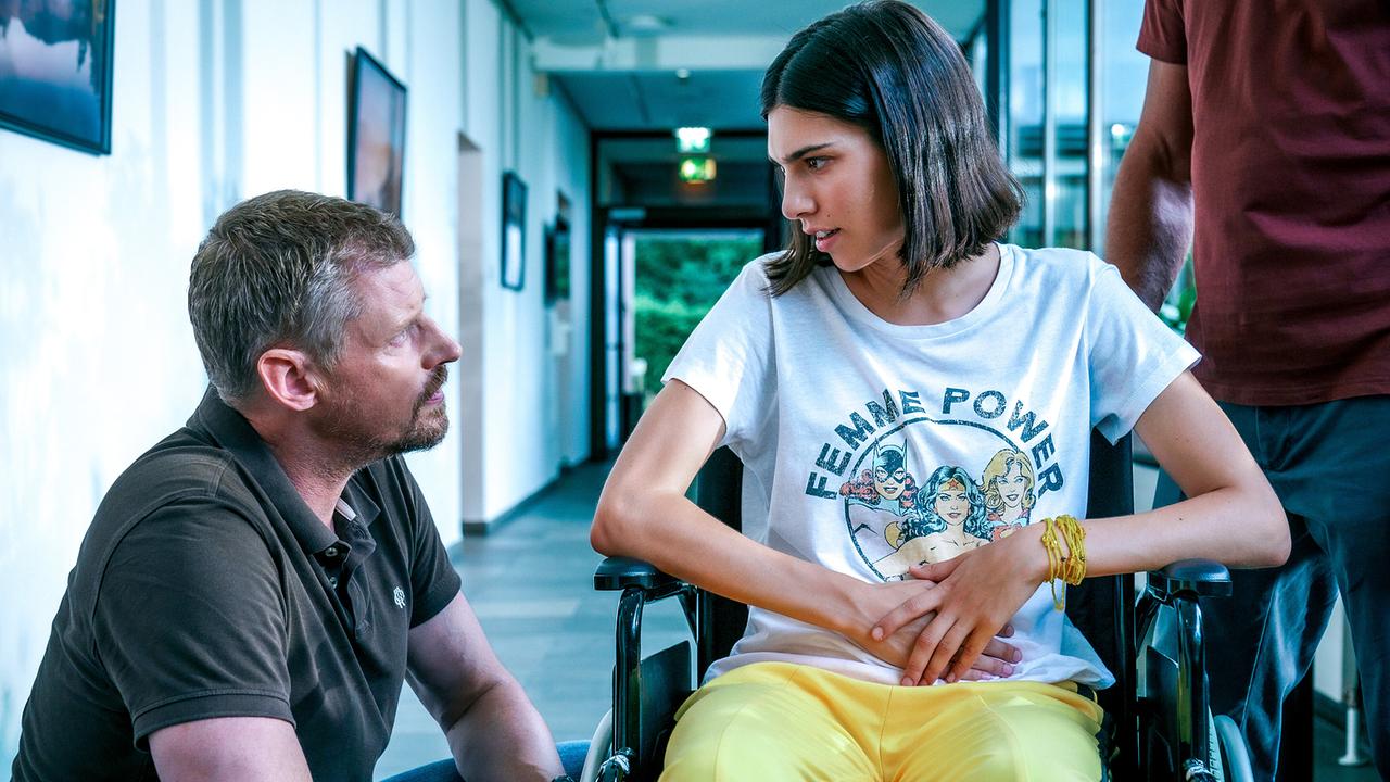 © ORF/Satel Film/Petro Domenigg Max Herzog hockerlt neben der jungen Laura, die in einem Rollstuhl sitzt und unterhält sich mit ihr in einem verglasen Gang.