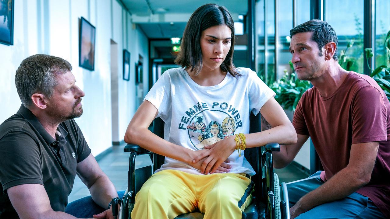 © ORF/Satel Film/Petro Domenigg In einem Gang mit Glasfassade sitzt das Mädchen Laura im Rollstuhl, flankiert Max Herzog und Klaus Lechner, die sich zu ihr hinunterbeugen.