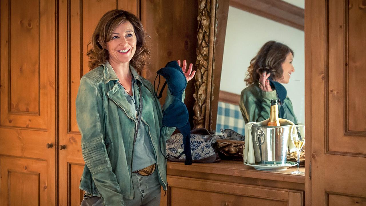  Nina Pokorny hält in einem rustikalen Wohnraum vor einem Spiegel  lächelnd einen BH hoch, neben ihr steht auf einer Kommode eine Kühlflasche mit Getränk.