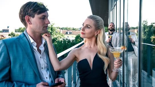 Silvia Schneider mit einem Cocktail in der Hand berührt das Kinn von Hubert Steiner auf der Terasse eines modernen Gebäudes