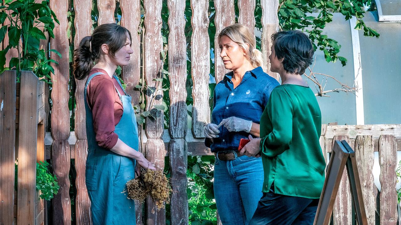 Anna Rieser mit ausgetrockneten Pflanzen in der Hand, Angelika Niedetzky und Katharina Stemberger unterhalten sich im Garten vor einem hohen Holzzaun
