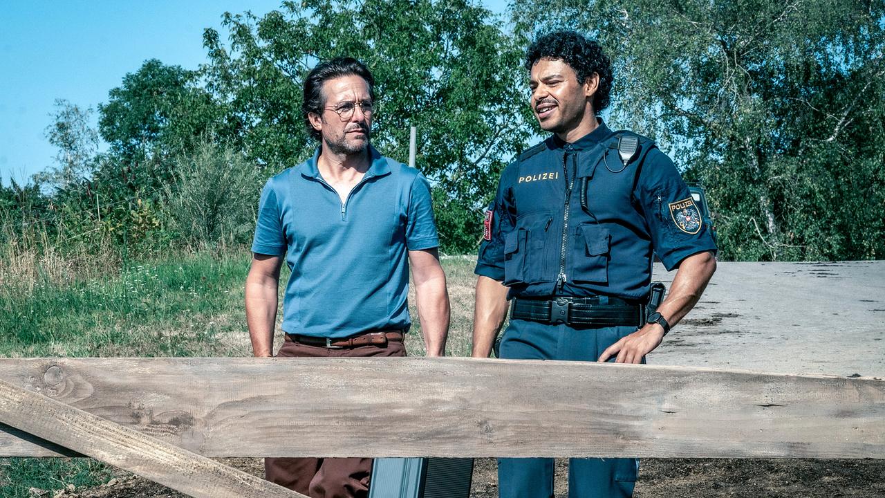 Alexander Pschill und Romeo Kaltenbrunner in Polizeiuniform stehen an einem Holzzaun im Grünen.