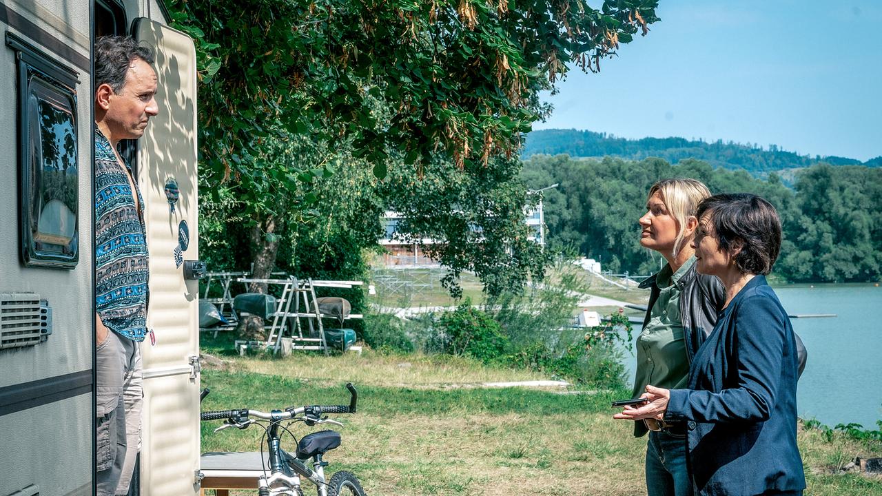 Angelika Niedetzky und Katharina Stemberger vor einem Wohnwagen an einem See, in dessen Eingang Stefko Hanushevsky steht.