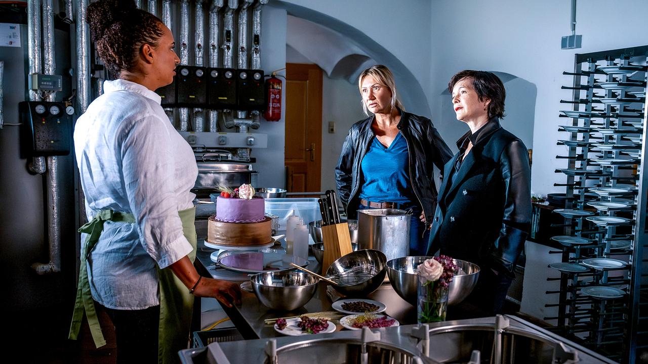 Angelika Niedetzky und Katharina Stemberger befragen Kira Lorenza Althaler in einer Berufskleidung für Gastronomie in einer Küche um einen Tisch mit einer dekorierten Torte und Backutensilien.