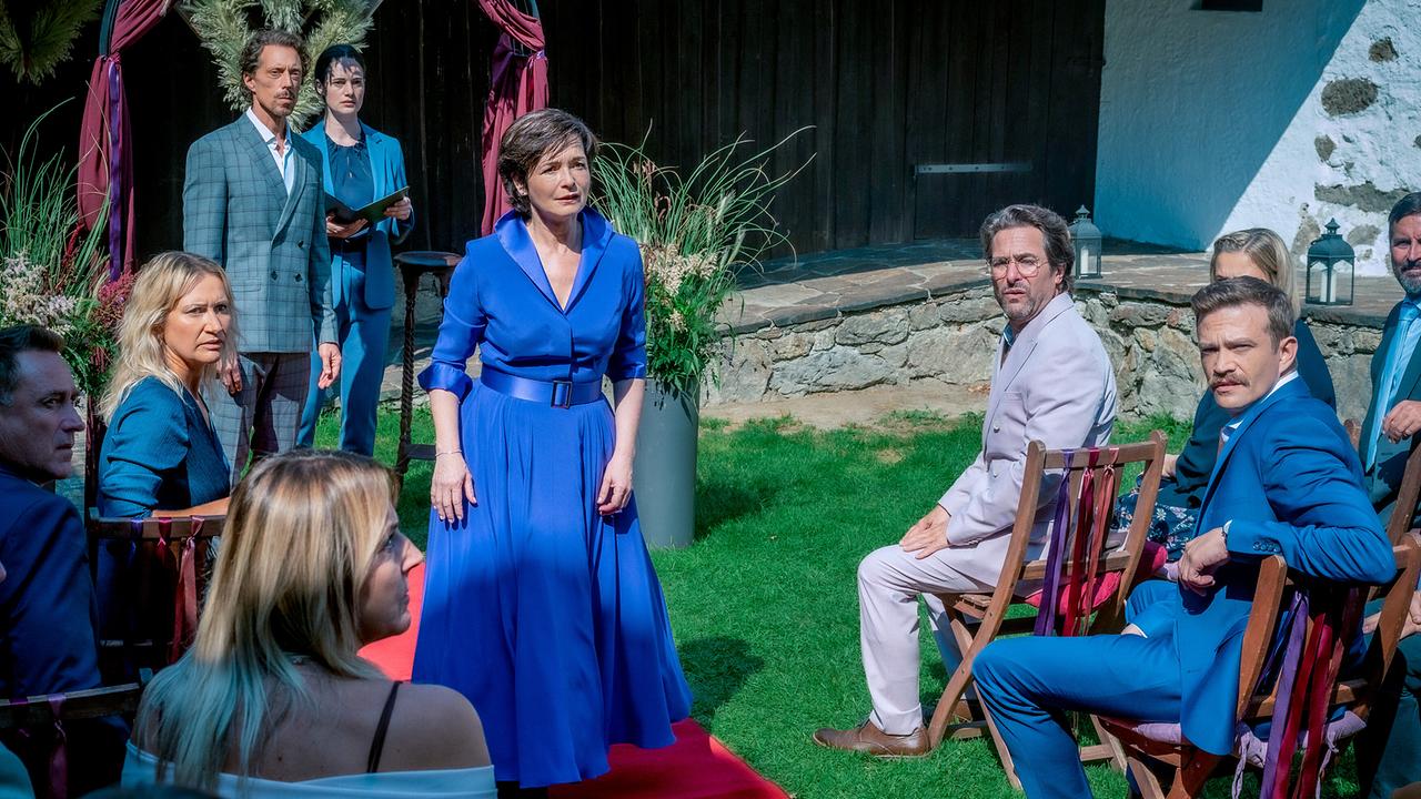 Katharina Stemberger in einem blauen Kleid mit Bräutigam Michael Pink, einer Standesbeamtin und die sitzende Gästen bei einer Hochzeitszeremonie im Freien auf einem roten Teppich drehen sich um und blicken auf etwas.