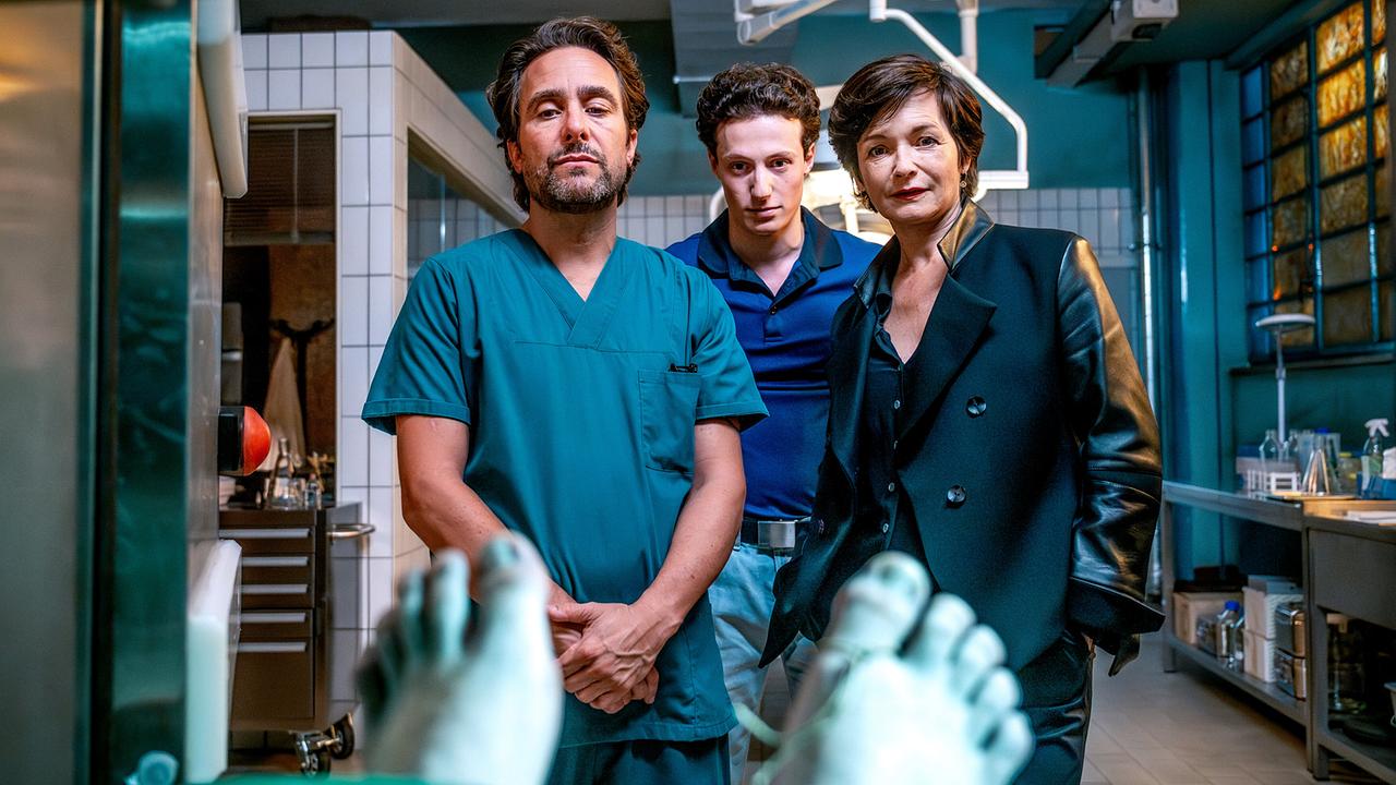 Alexander Pschill, Damyan Andreev und Katharina Stemberger stehen in einem Obduktionsraum nebeneinander und  blicken auf einen bedeckten Körper auf einem Seziertisch.