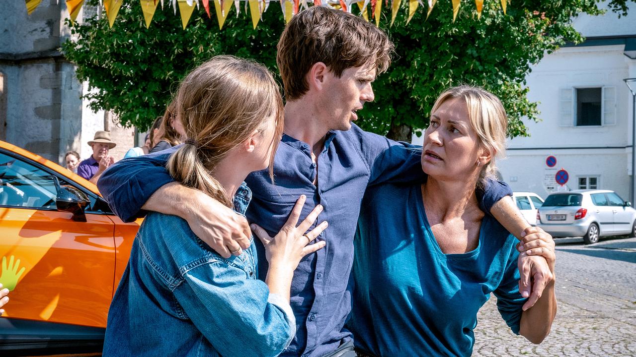 Nikola Jaritz-Rudle und Angelika Niedetzky stützen Lukas Strasser unter einer gelben Girlande vor einem orangefarbenen Auto.