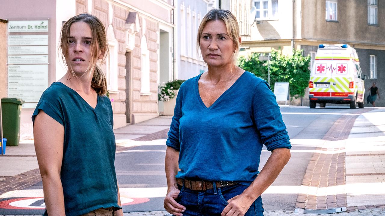Nikola Jaritz-Rudle und Angelika Niedetzky stehen ernst blickend auf einer Straße, im Hintergrund ist ein Rettungswagen zu sehen.