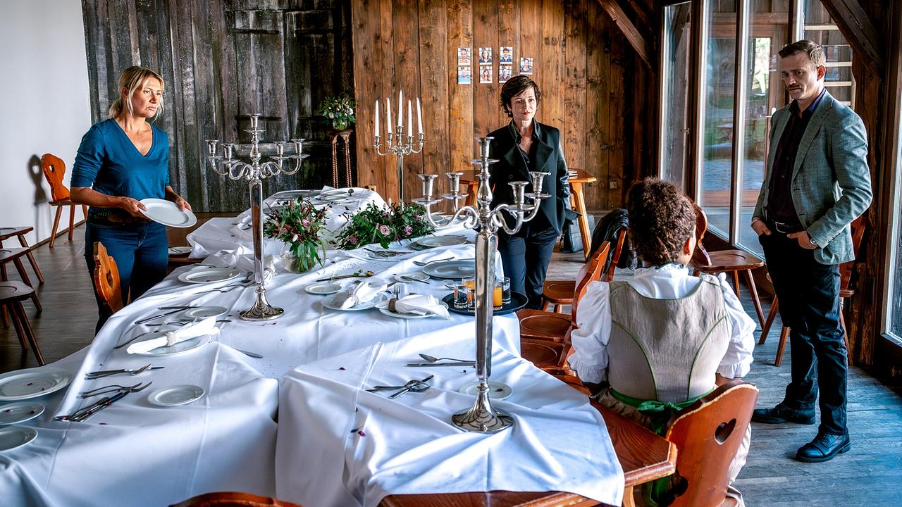 Angelika Niedetzky, Katharina Stemberger und Michael Steinocher sprechen mit der sitzenden Kira Lorenza Althaler  in einem rustikalen Raum mit einem langen, festlich gedeckten Tisch und Kerzenleuchtern.