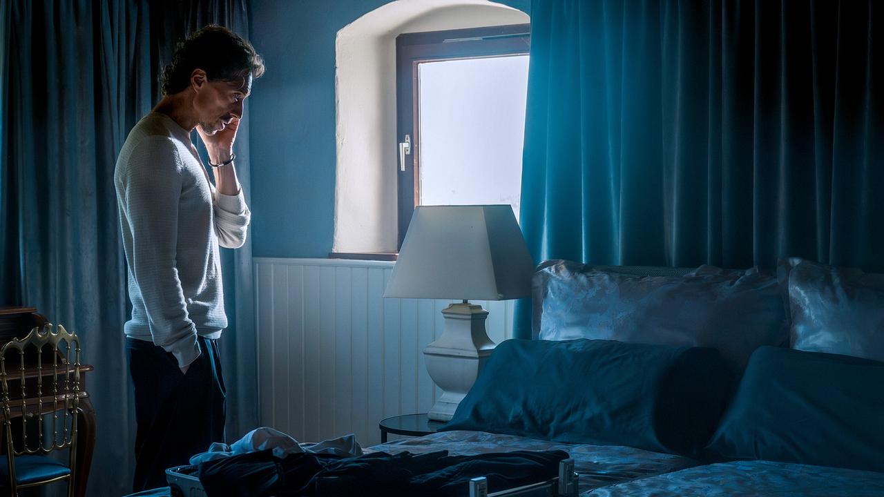 Michael Pink steht nachdenklich in einem blauen, abgedunkelten Schlafzimmer neben einem geöffneten Koffer und telefoniert.
