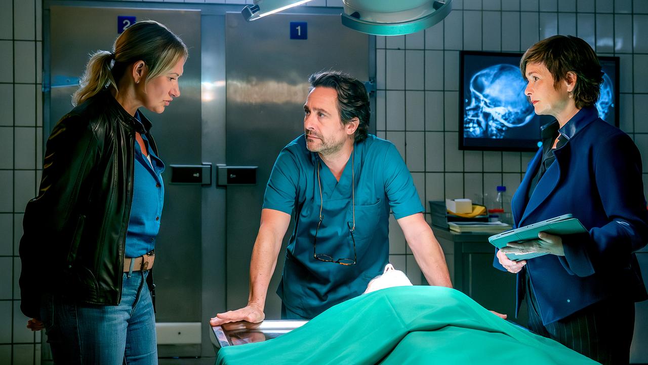 In einem Obduktionsraum stehen Biggi Obergschwendter und Joe Haizinger mit Dr. Richie Vitek im Kasack um einen Tisch mit einem abgedeckten Leichnam.