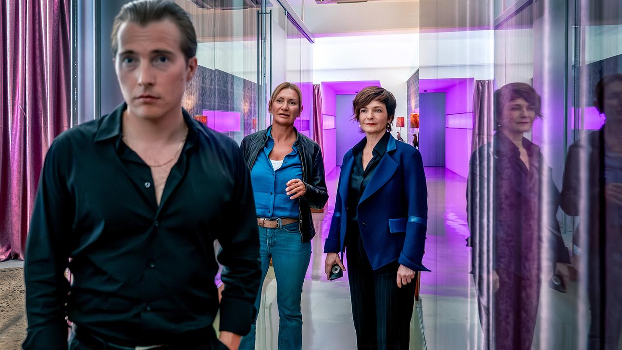 Samy Rahs, ein junger Mann, steht in einem modern beleuchteten Raum mit dem Rücken zu Biggi und Joe.