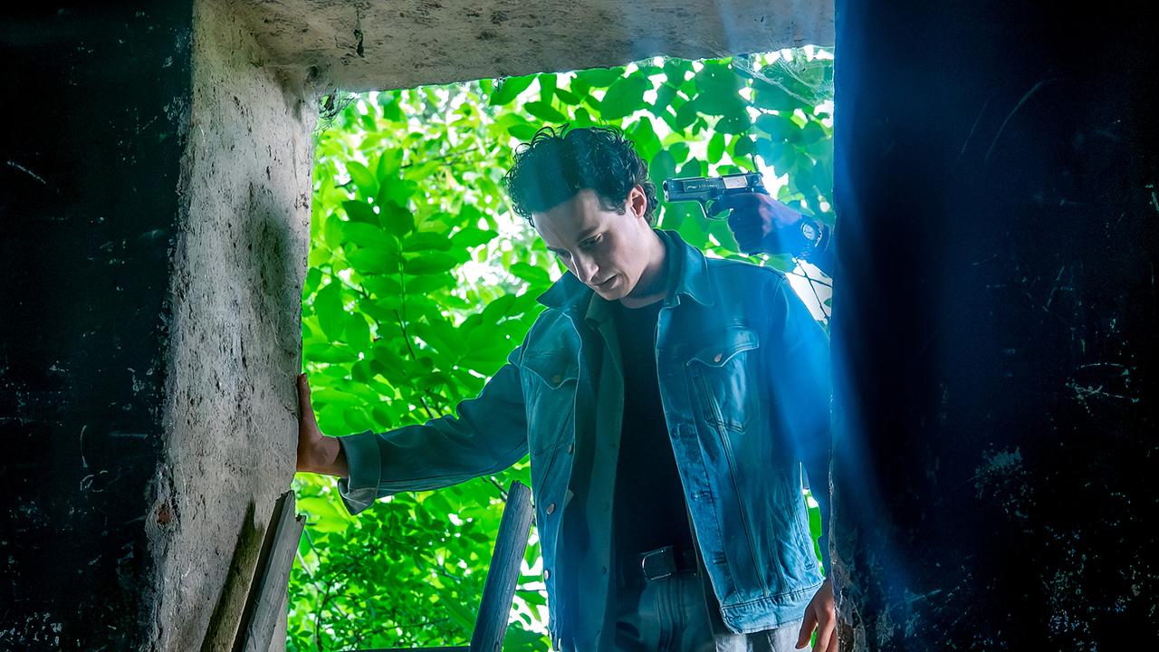 Aleks Malenov steht im Grünen in einer dunklen Maueröffnung, während eine unbekannte Person ihm eine Pistole an den Kopf hält.