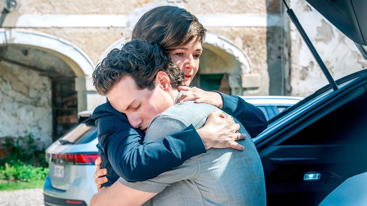 Joe umarmt Aleks Malenov tröstend neben einem geöffneten Kofferraum vor einem alten Gebäude.