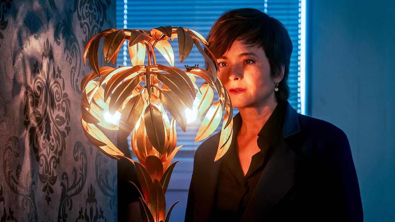 Joe Haizinger betrachtet besorgt eine goldene kunstvolle, blattförmige Stehlampe in einem verdunkelten Zimmer.