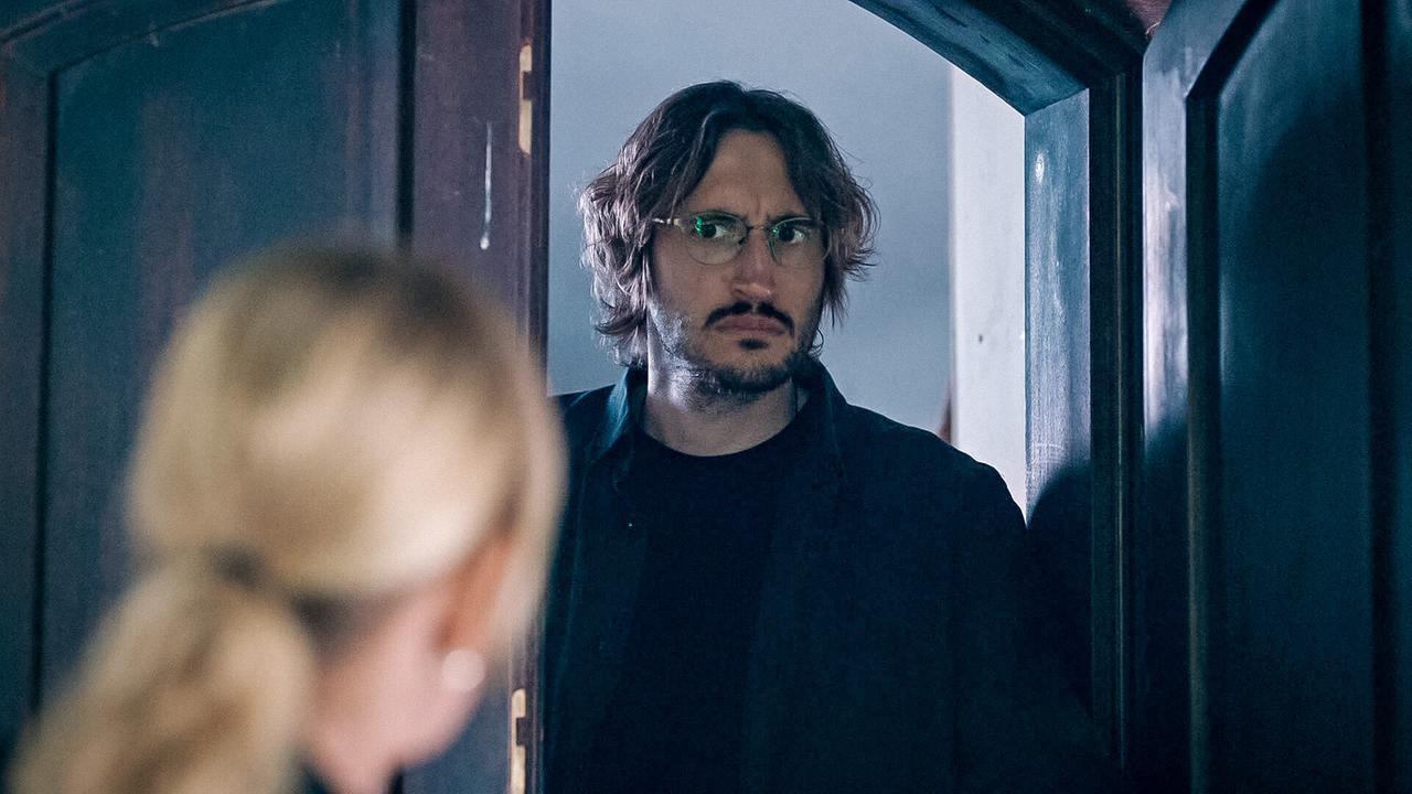 Paul Bruckner, ein bärtiger Mann mit Brille steht ernst blickend in einer geöffneten Tür, während im Vordergrund Biggi Obergschwendter unscharf zu sehen ist.