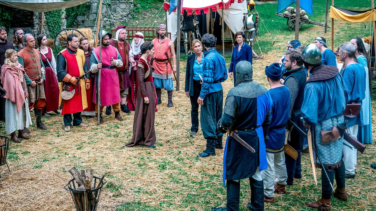 In einem mittelalterlichen Lager treffen die modern gekleideten Biggi Obergschwendter und Joe Haizinger auf eine Gruppe kostümierter Ritter und mitteralterlich gekleidete Dorfbewohner:innen.